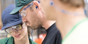 Trois employés portant des lunettes de protection et des bouchons d'oreilles discutent dans une usine, mettant l'accent sur la sécurité au travail.