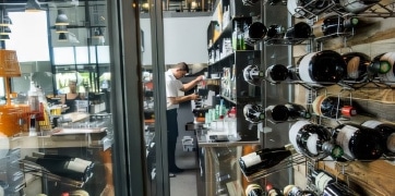 Un employé gère les stocks de bouteilles de vin dans un bar moderne. Les bouteilles sont rangées sur des étagères murales métalliques.